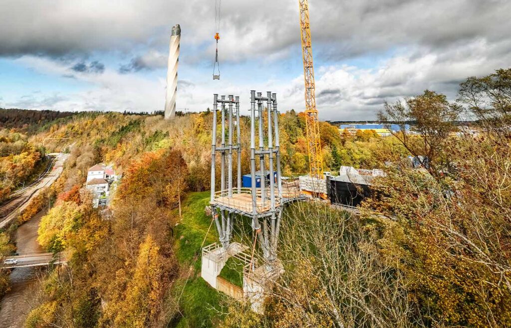 Übersicht auf eine Baustelle mit Kran zwischen Wald im Herbst, im Hintergrund ein futuristischer Turm