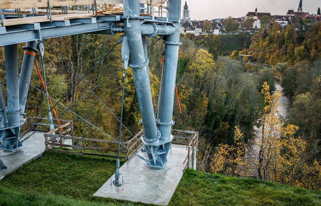 Nahaufnahme einer Stahlkonstruktion am Hang vor einem Fluss und einer Stadt