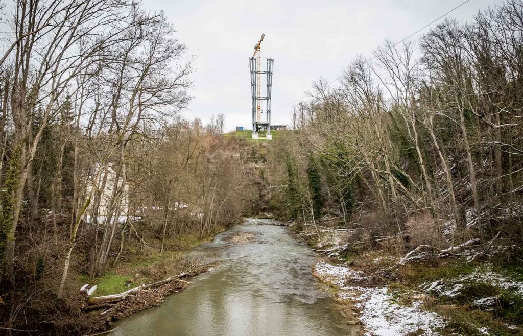 Fluss im Vordergrund, links und rechts Bäume und im Hintergrund der Pylon der Neckarline