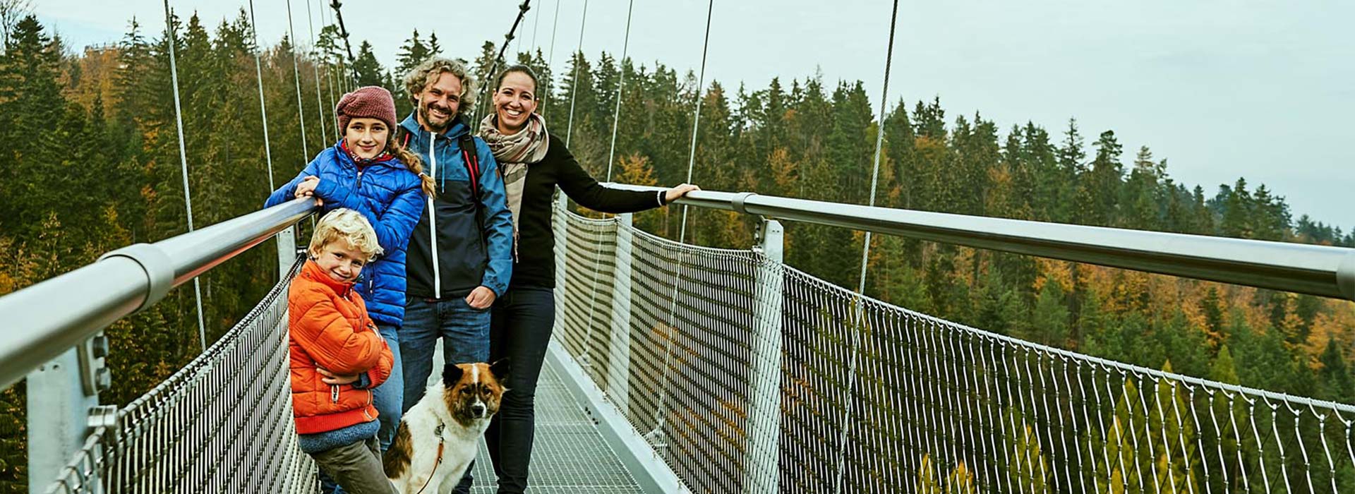 Eine europäische Familie auf einer Hängeseilbrücke im Herbst mit Jacken und einem Hund, dahinter Tannenbäume