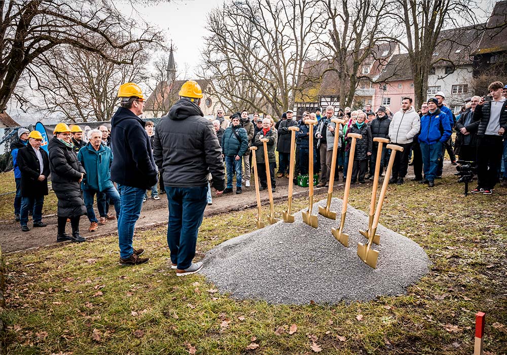 Ansicht auf eine Menschengruppe mit zwei Personen im Vordergrund neben acht Schaufeln in Kies