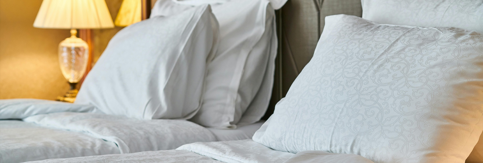Nahaufnahme eines Hotel-Doppelbetts mit frischer Bettwäsche mit leichtem Muster und einer historischen Nachttischlampe
