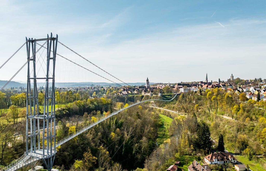 Abbildung einer Hängebrücke mit Altstadt im Hintergrund.