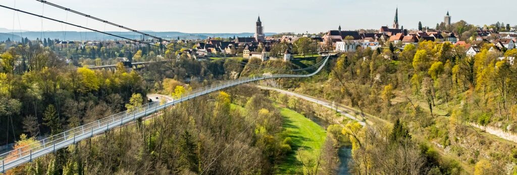 Abbildung einer Hängebrücke mit Altstadt im Hintergrund.